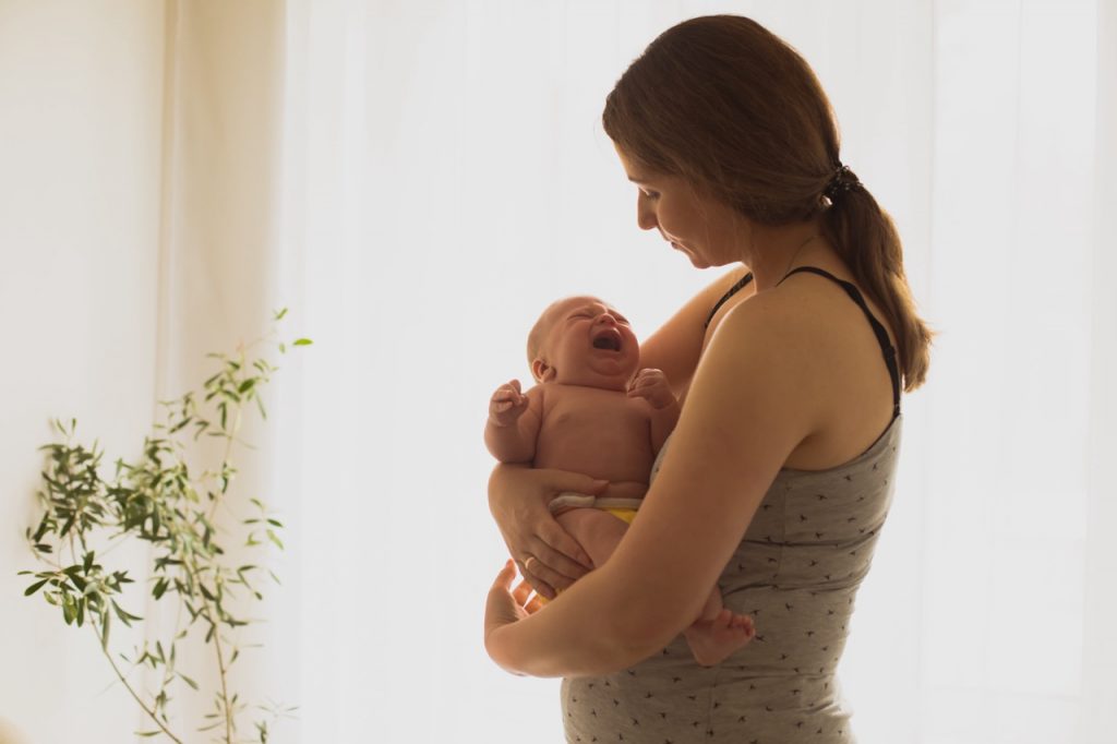 Young unemotional mother holding crying newborn in her arms. Woman in postpartum depression has no strenght to calm down her crying baby. Lonely exhausted mother at home.