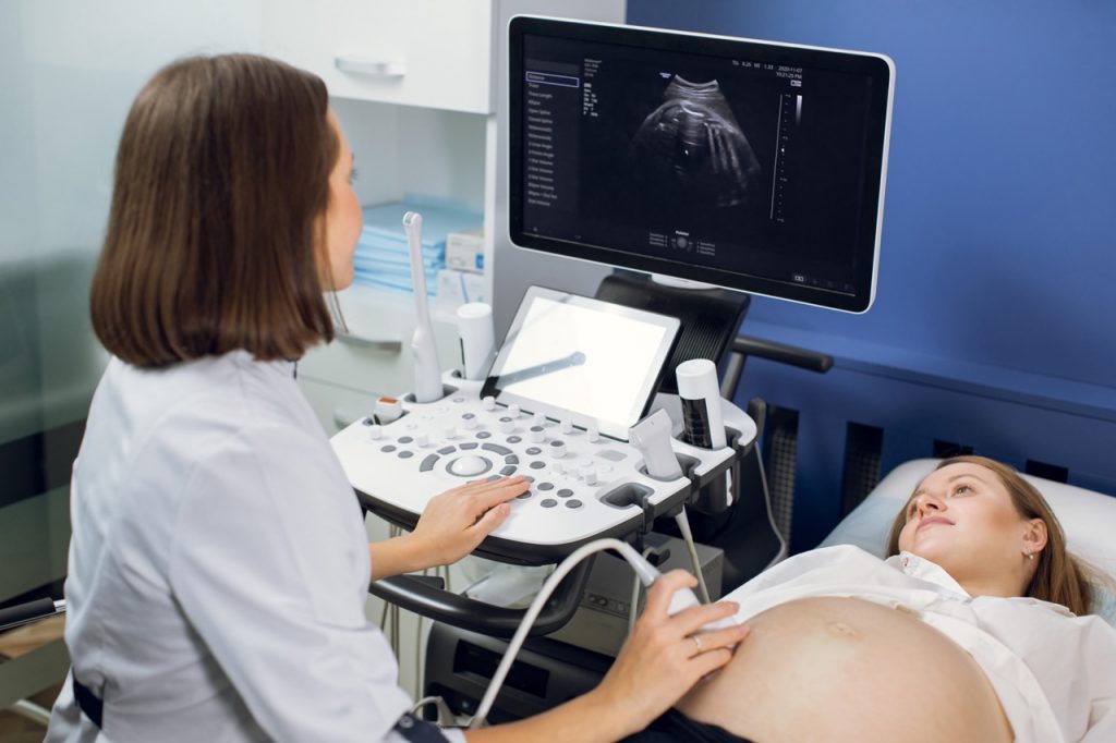 Happy pregnancy time, ultrasound concept. Pretty pregnant woman doing ultrasound scan in the prenatal clinic, worried about health of her future baby. Female doctor looking at the screen.